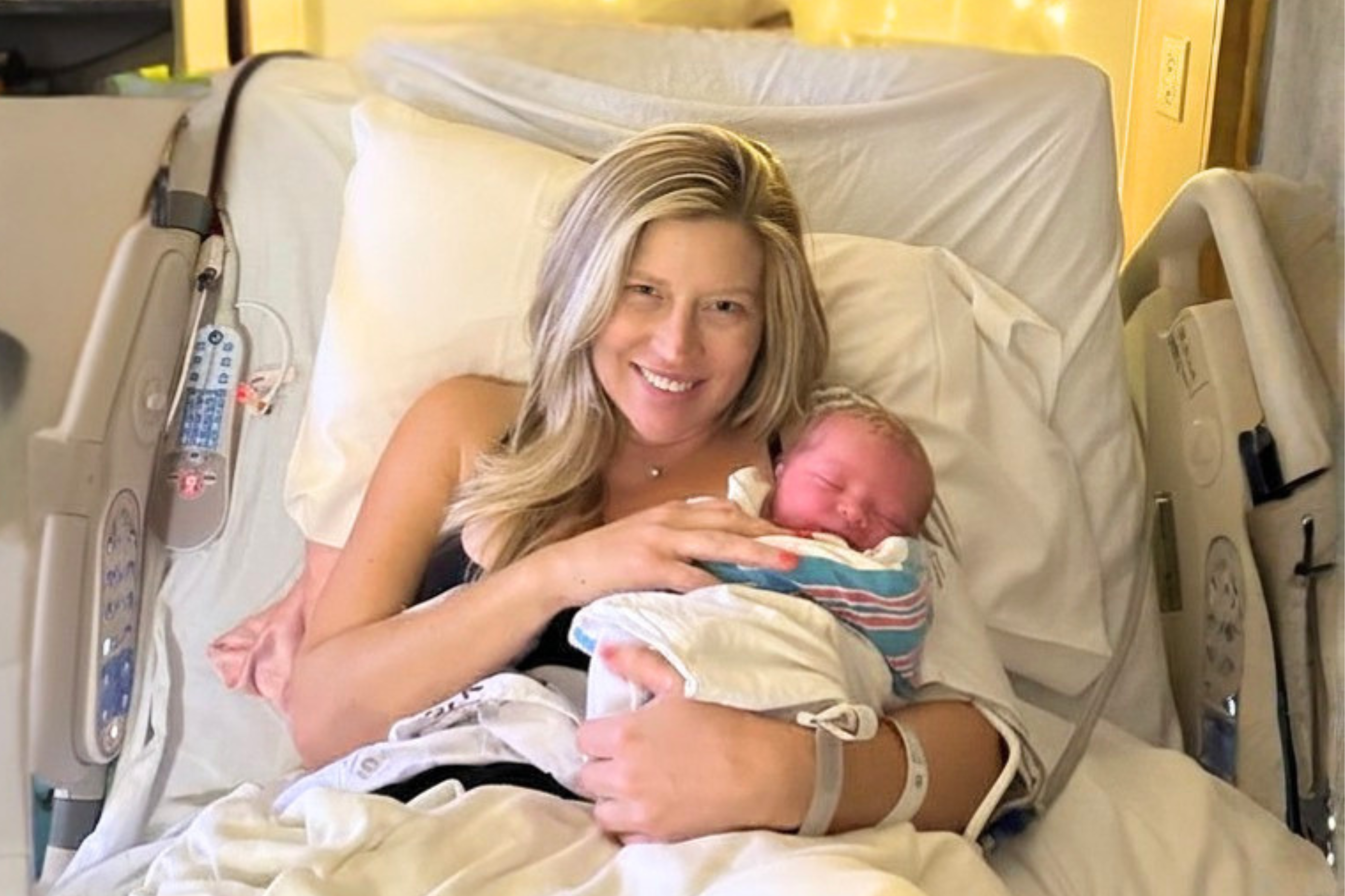 New mother smiling while holding her newborn baby in a hospital bed — postpartum recovery begins here
